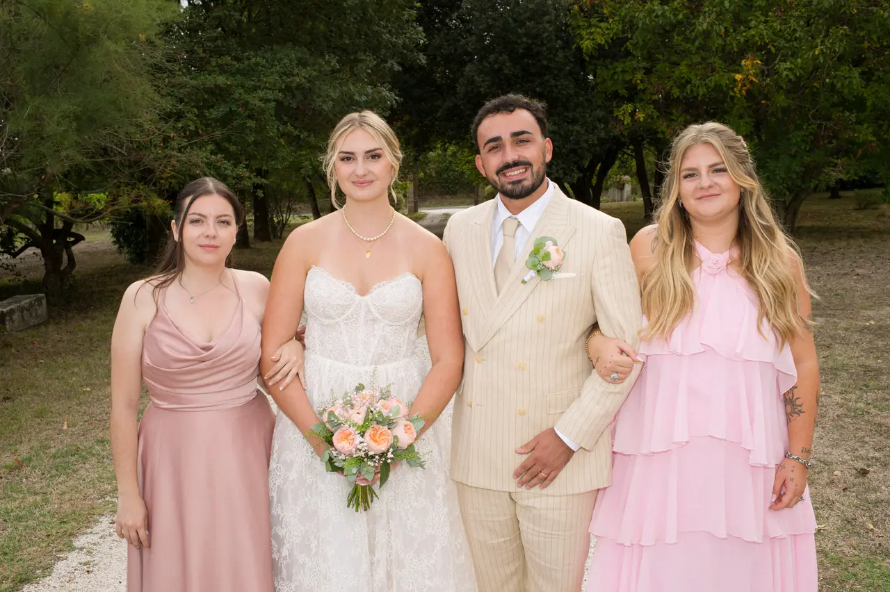 mariés bras dessus bras dessous entre deux femmes en robes roses, parc arboré