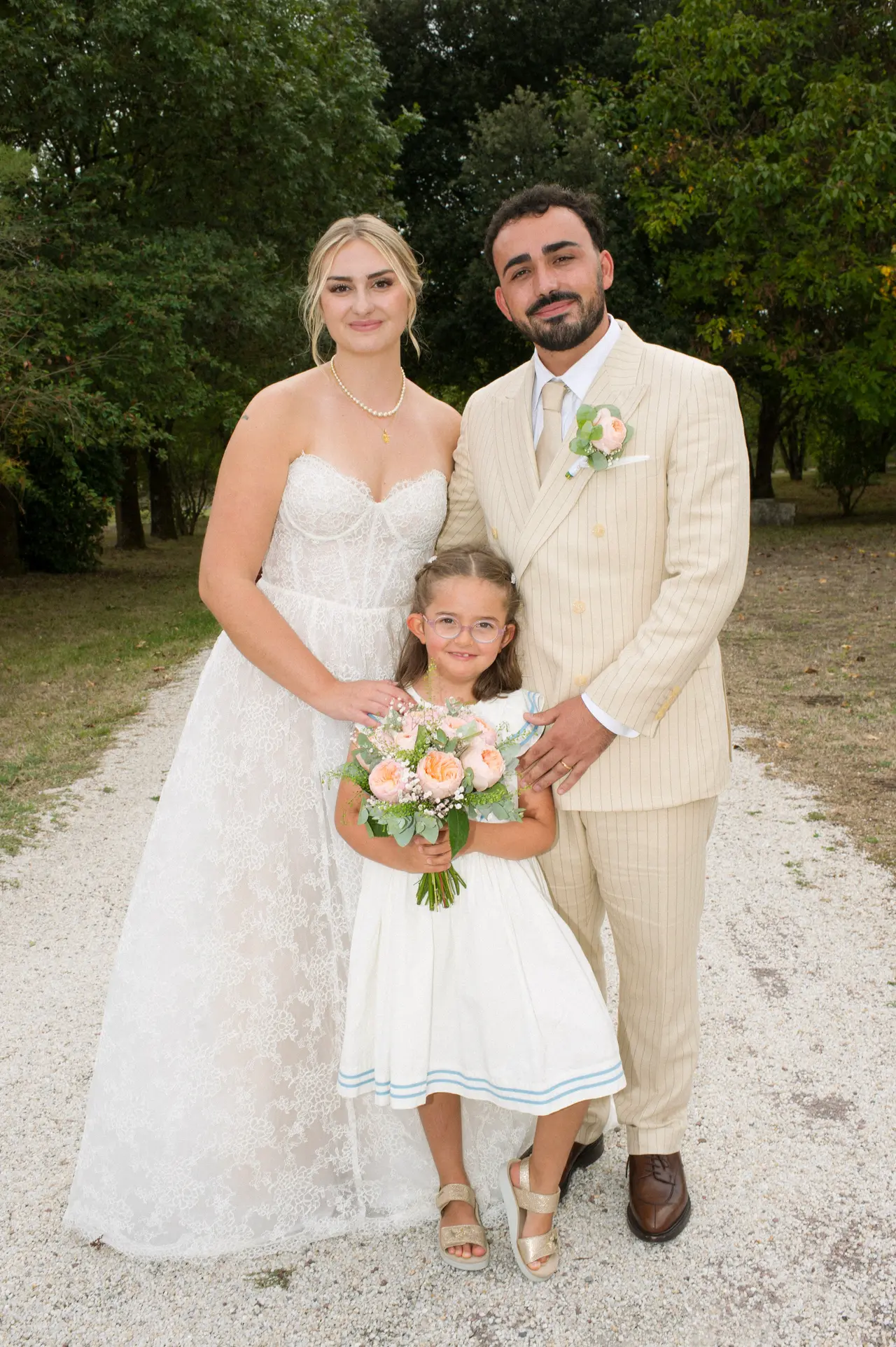les mariés debout avec une fillette en robe blanche tenant le bouquet, allée de gravier