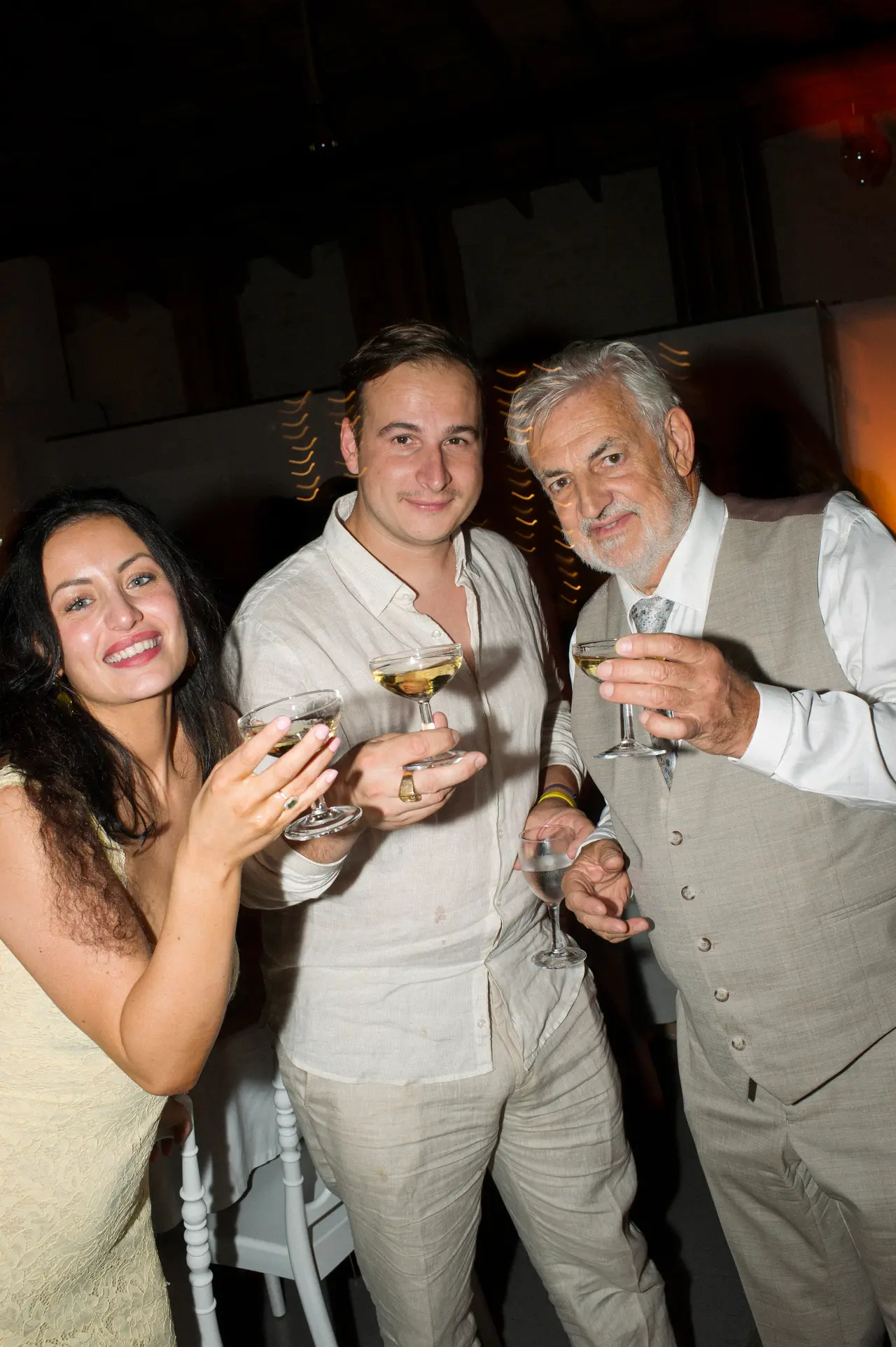 Trois invités levant des coupes, robe crème, chemise lin et gilet beige, de nuit